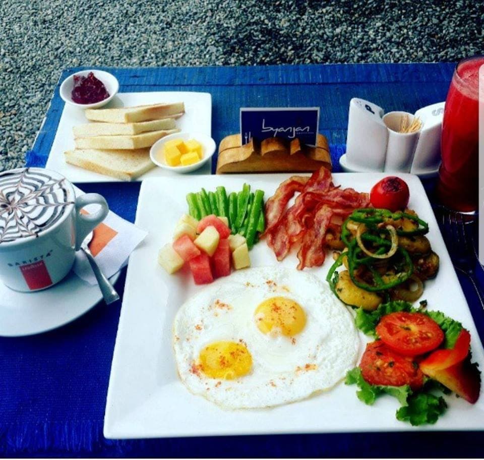 A full breakfast on a blue mat: fried eggs, bacon, toast, fruit and a cappuccino.