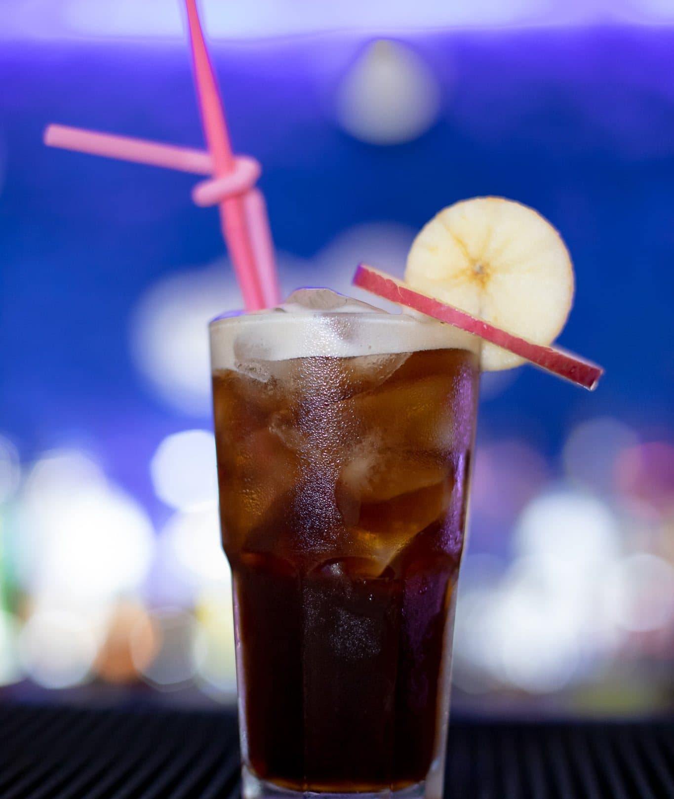 An iced cooler with an apple slice and pink straws, blue bar lights blurred behind.