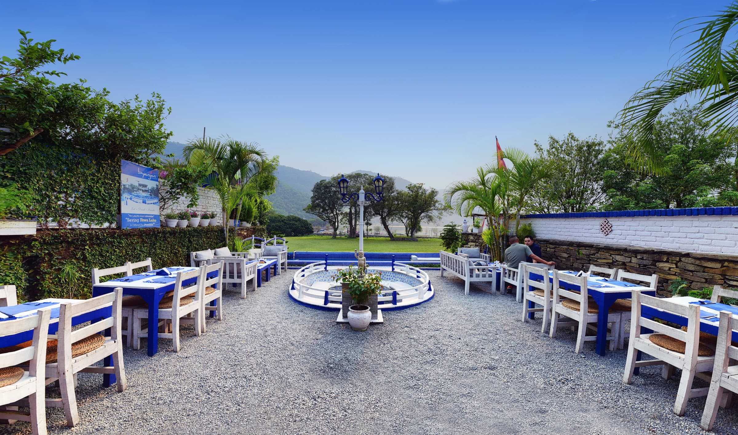 Byanjan’s patio by day: blue tables and white chairs around the sunken seating, palms, Phewa Lake and the hills beyond.